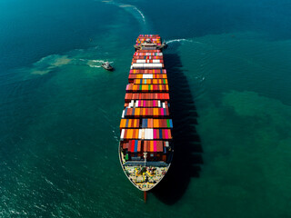 Aerial front view of long container ship sailing in deep blue ocean with tugboats