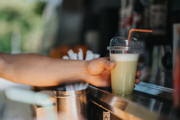 Close-up of a person serving a refreshing beverage in a transparent plastic cup with a straw, depicting a warm day and outdoor activity at a modern mobile drink stand.