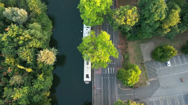 An aerial view shows a serene river bordered by lush green trees. A white houseboat is docked along the bank, adjacent to a road with a pedestrian crossing.