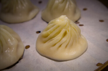 A close-up of a single Xiao Long Bao (soup dumpling) with its characteristic pleated top, sitting on a parchment liner in a steamer basket. 
