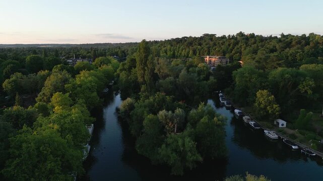 Aerial view of a serene river winding through lush green trees, with boats docked along the bank and a large building nestled in the distant foliage.