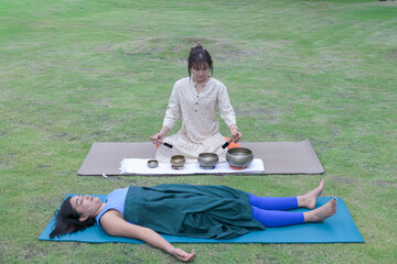 Woman performing sound healing with Tibetan singing bowls during relaxation yoga session outdoors.