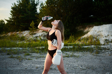 Young woman in black bikini and boxing gloves drinking water from a bottle in a natural environment