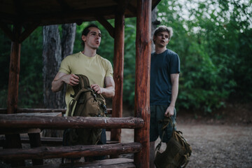 Two men with backpacks pause at a rustic wooden shelter in a lush forest, ready for an outdoor hike. Casual gear, camaraderie, and nature set the mood for outdoor adventure.