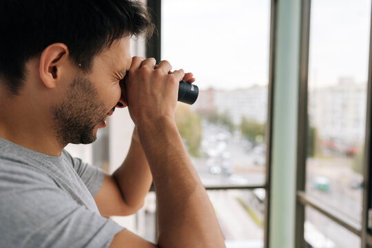 Curious young adult man holding monocular, looking closely out of window at urban street, engaging in observation, surveillance and spying while monitoring surroundings from apartment or home.