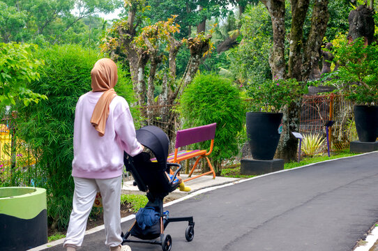A woman pushes a baby stroller along a quiet garden pathway, enjoying a peaceful walk surrounded by greenery in a calm outdoor park setting. - Powered by Adobe