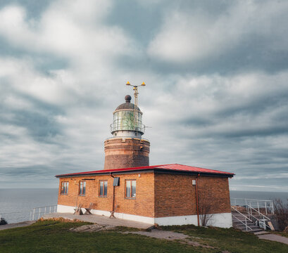 A wide, square-format photograph of the historic Kullen Lighthouse (Kullens Fyr) situated on the rugged cliffs of Kullaberg, Sweden. The brick building and cylindrical stone tower with its copper dome