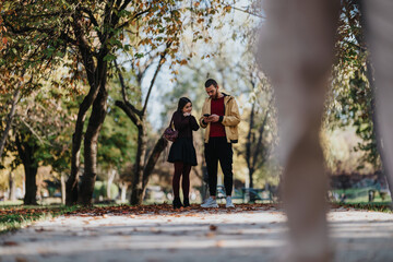 A couple stands on a sunlit park path during fall, sharing a moment as they check a smartphone. Fallen leaves, trees, and a peaceful atmosphere create a warm, candid scene.