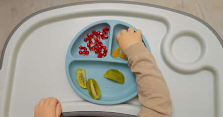Little boy is happily eating a variety of fruits at home. He explores different textures and tastes while sitting in his high chair, demonstrating a love for healthy snacks.