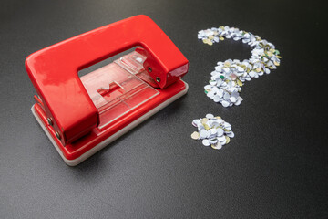 Red hole puncher with paper confetti arranged as a question mark symbol on black background. Concept of business problem, administrative confusion, or office work inquiries.