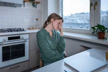 Exhausted woman covering face with hand feels burnout, drained, sits at table in front of closed laptop. Tired female with mental fatigue, emotional pressure, anxiety caused by workload, life problems