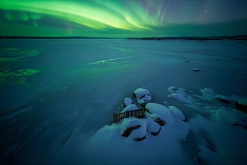 Bright green northern lights fill the night sky over a snowy field. Old wooden ruins lie in the snow, creating a contrast against the bright aurora above.
