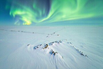 Bright green northern lights fill the night sky over a snowy field. Old wooden ruins lie in the snow, creating a contrast against the bright aurora above.