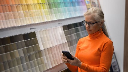 Mature woman looks at her phone while standing in front of paint color samples in a large hardware store during a shopping trip