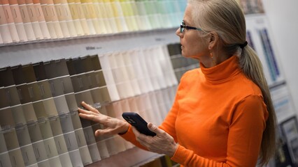 Mature woman studies paint color samples in a large hardware store while holding her phone and planning for home renovation