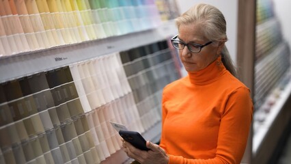 Mature woman stands in a hardware store looking at paint samples while checking her phone for home renovation ideas