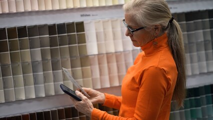 Mature woman looks at paint samples on a display while using her phone for shopping during a visit to the hardware store