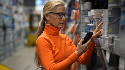 Mature woman shops in a large hardware store, checking her phone while looking at products for home...
