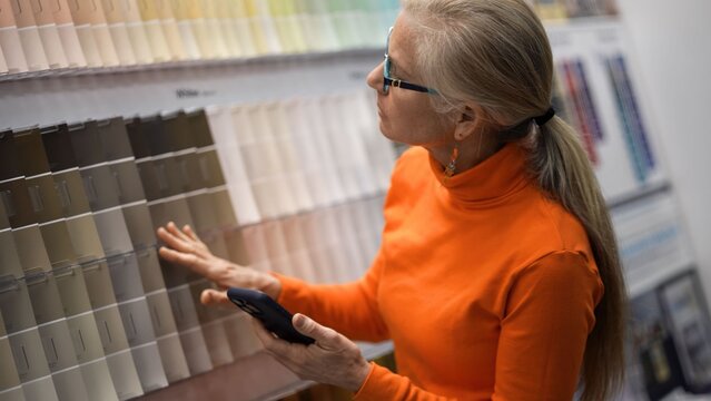 Mature woman looks at color samples in a hardware store while planning for home renovation projects with her phone in hand