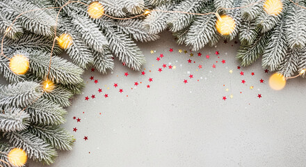 Top-down view of snowy fir branch and light garland. Red star confetti scattered on gray backdrop. It represents festive holiday, celebration season