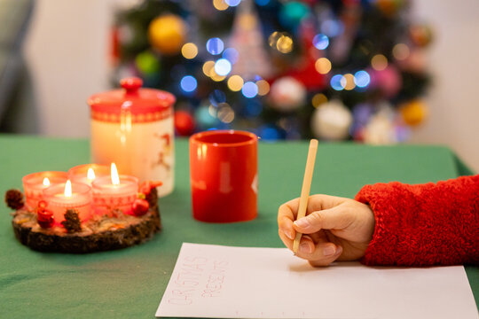 Mano femminile che scrive su un foglio bianco la lista dei regali di Natale, su un tavolo natalizio, alle spalle le luci dell'albero di Natale