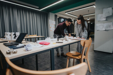 Two students collaborate in a modern design studio, reviewing blueprints, building models, and planning a project. Laptops, sketches, and materials are spread across the table.