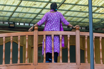 Back view of an elegant Bangladeshi woman in a traditional purple Salwar Kameez, standing peacefully beside a wooden railing. South Asian culture, fashion, and lifestyle concept.