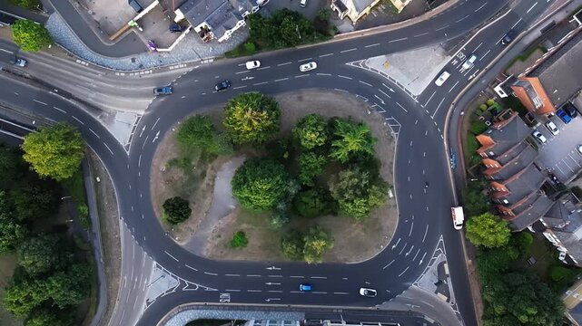 An aerial drone view shows a busy roundabout with cars navigating, surrounded by trees and residential housing.