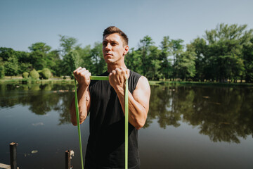 Man performing fitness exercises outdoors using a resistance band near a lake. Surrounded by lush greenery, he engages in physical training while enjoying the tranquil environment of nature.