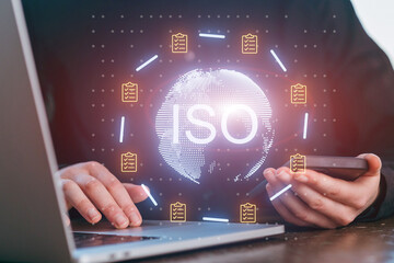 Person holding smartphone and working on laptop with data on ISO standards displayed on screen