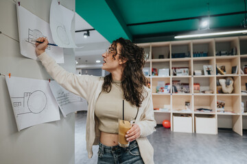 A woman in casual attire pins architectural drawings on a wall in a bright collaboration center, holding an iced coffee and watching her sketches.