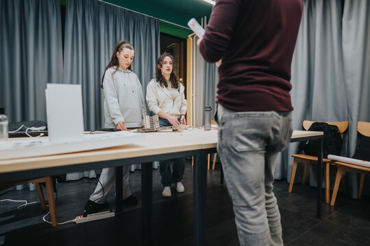 Two women stand at a table in a collaboration center as a man in the foreground presents. They discuss materials for a project in a focused, collaborative workshop setting.
