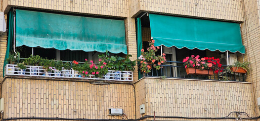 Two balconies in Granada, Spain, with green awnings and vibrant potted flowers on brick facade, showing a cozy urban lifestyle. residential