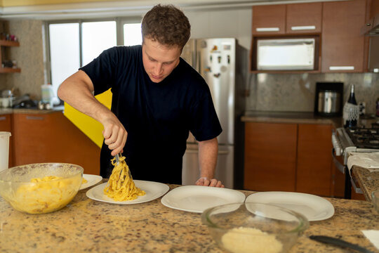 A young Italian chef is serving pasta carbonara onto a plate in a kitchen.
