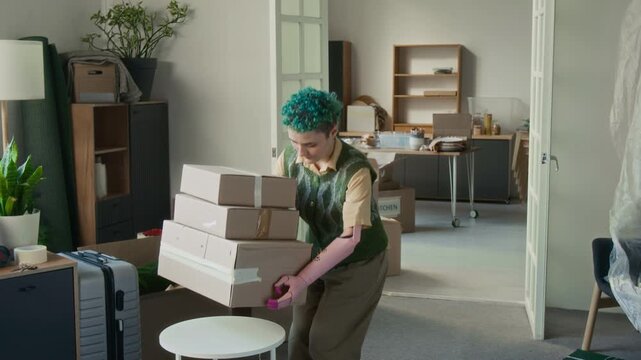 Gen Z girl with prosthetic arm carrying three cardboard boxes to living room and putting them on round coffee table while moving in