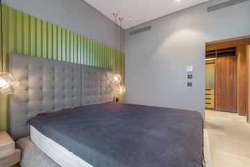 The scene shows a modern bedroom featuring a large bed with a dark cover. Unique light fixtures hang above, adding to the design. The walls have a mix of colors and textures, creating visual interest.