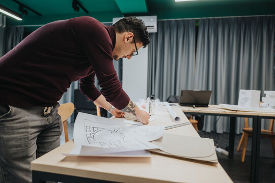 A man wearing glasses leans over a long table, reviewing blueprints and drafting plans in a bright collaboration center. Laptops and architectural drawings spread across the workspace. - Powered by Adobe
