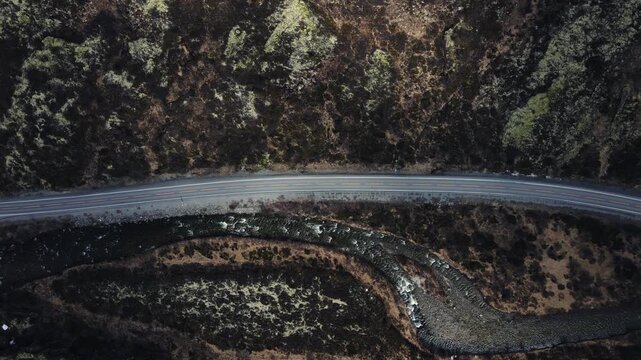 Vertical top-down aerial drone shot of an asphalt mountain road running parallel to a flowing river through rocky mossy terrain in the Norwegian wilderness.