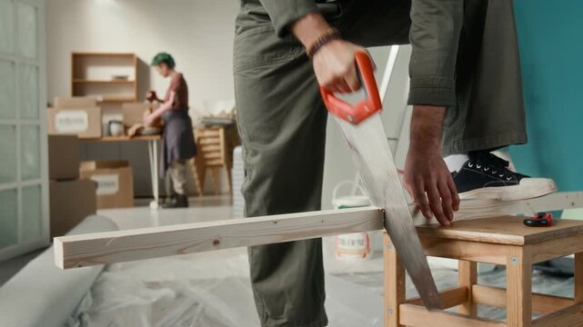 Unrecognisable man wearing jumpsuit using handsaw to cut plank while standing on wooden stool with one foot, girlfriend unpacking kitchen items in background