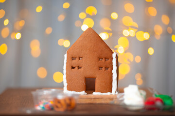 Horizontal photo. Gingerbread house without roof with white icing decor on wood cut board table. Golden garland, bokeh lights on light curtain. Concept of Happy New Year, sweet food, Merry Christmas