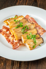 Fried eryngii mushrooms served with crispy bacon slices on white plate