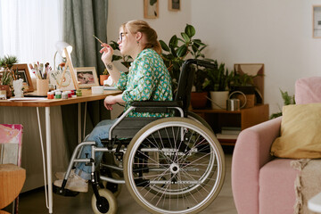 Caucasian young adult woman with disability sitting in wheelchair working at desk holding paintbrush near art supplies surrounded by houseplants and framed photos 
