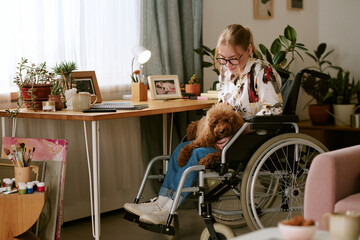 Caucasian young adult woman with disability sitting in wheelchair holding brown poodle dog in home...