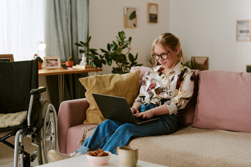 Caucasian young adult woman with disability sitting on sofa using laptop, smiling while working or...