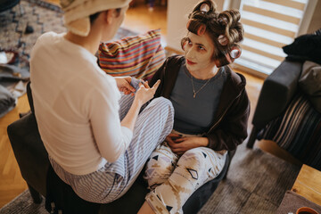 Two women relax at home while one wears curlers and a facial mask, chatting in a cozy, intimate...