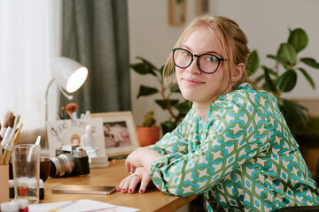 Portrait of Caucasian young adult woman with blond hair and glasses sitting at desk turning toward camera smiling, surrounded by art supplies and camera, home environment visible