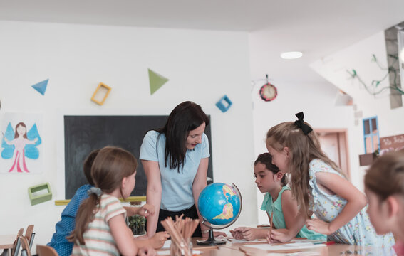 Female teacher with kids in geography class looking at globe. Side view of group of diverse happy school kids with globe in classroom at school.