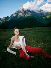 Naklejka premium Adventure in a meadow with towering mountains, a smiling woman sits on grass after a workout, bright red pants, white top, towel over shoulder, natural scenery