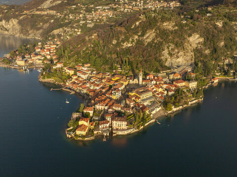 Aerial view of the sun-kissed Varenna, with its cluster of red-roofed buildings nestled along the serene waters of Lake Como, shadowed by the rugged mountains, Varenna, Lombardia, Italy.