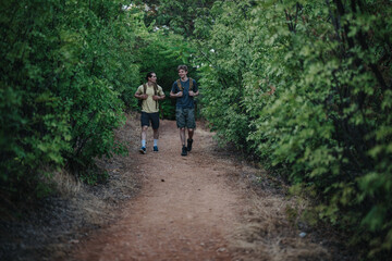 Fototapeta premium Hikers walk along a dirt forest trail, chatting and enjoying nature. They carry backpacks and wear casual outdoor clothing, capturing a moment of friendship, fitness, and adventure in lush greenery.